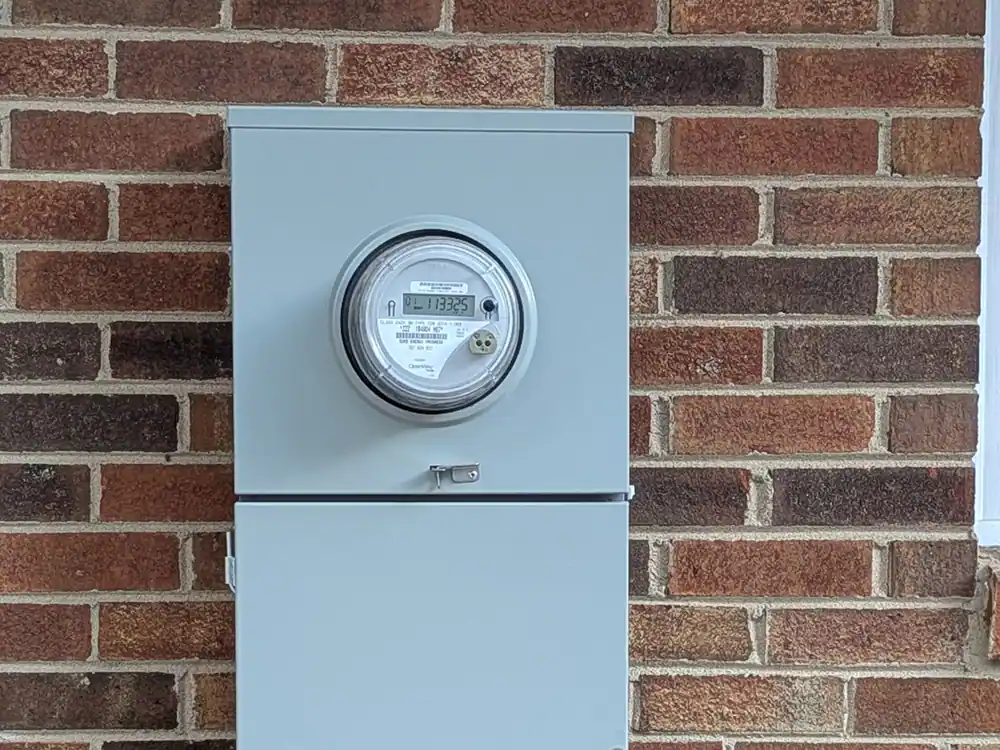A digital electricity meter mounted on a brick exterior wall, with a gray metal enclosure directly below it, in Glenwood-Brooklyn, Raleigh