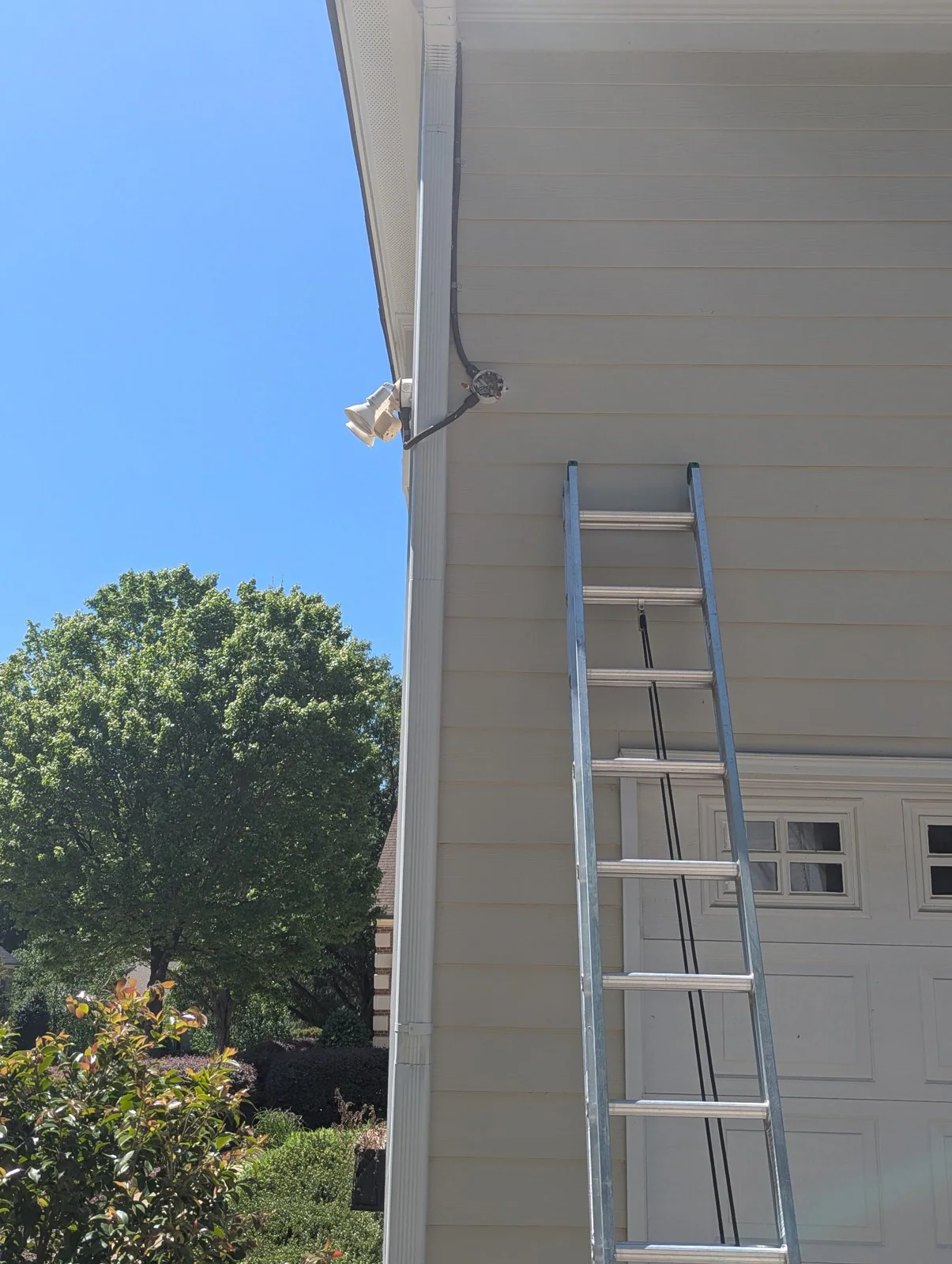 A ladder leaning against a house beneath an outdoor light fixture with wiring above it, in Budleigh, Raleigh