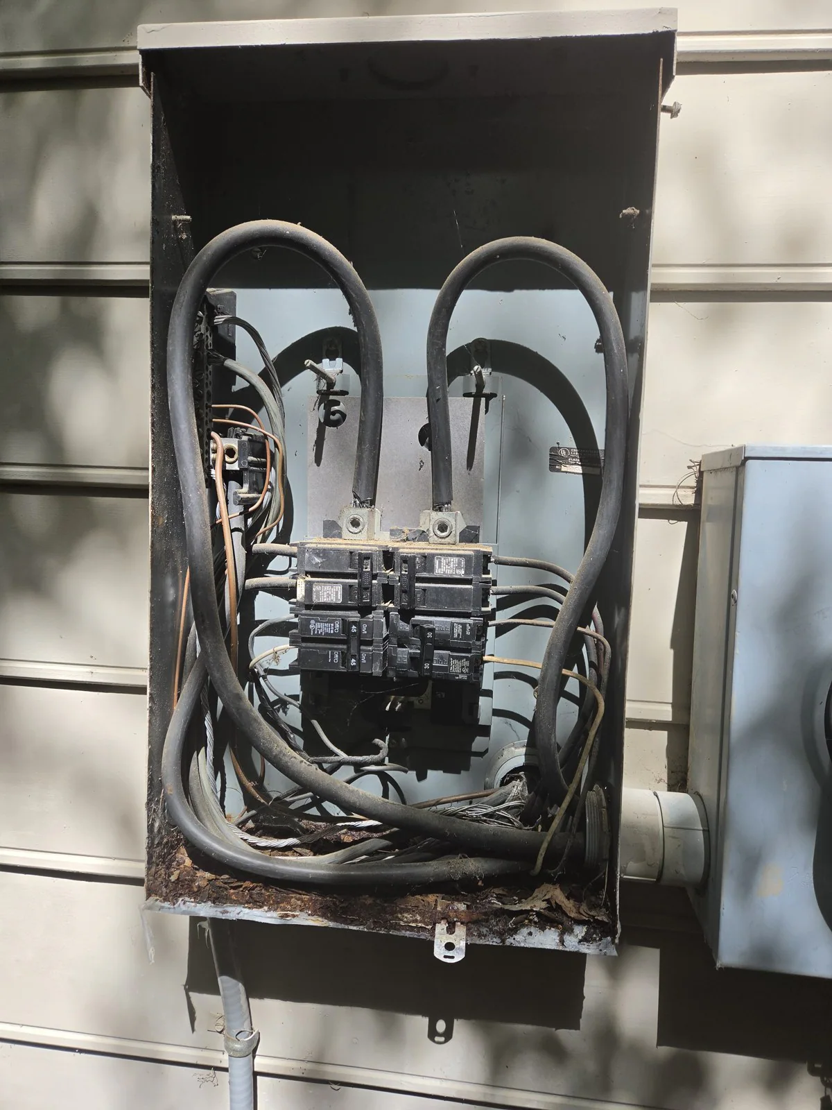 An open electrical panel with tangled wires and a rusted bottom section, in Oakwood, Raleigh
